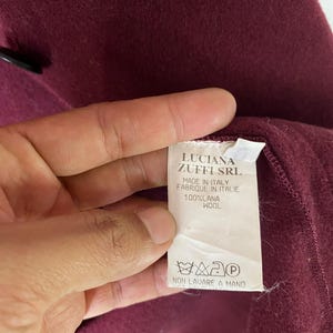Puede incluir: Una etiqueta blanca con texto negro que dice "LUCIANA ZUFFI SRL MADE IN ITALY FABRIQUE IN ITALIE 100%LANA WOOL" y un s&iacute;mbolo de instrucciones de lavado. La etiqueta est&aacute; unida a una tela burdeos.