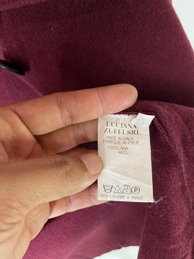 Puede incluir: Una etiqueta blanca con texto negro que dice "LUCIANA ZUFFI SRL MADE IN ITALY FABRIQUE IN ITALIE 100%LANA WOOL" y un s&iacute;mbolo de instrucciones de lavado. La etiqueta est&aacute; unida a una tela burdeos.