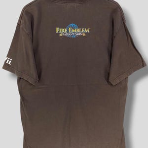 May include: Brown t-shirt with the text "Fire Emblem Radiant Dawn" in gold and blue. The shirt has a crew neck and short sleeves. The Wii logo is visible on the left sleeve.