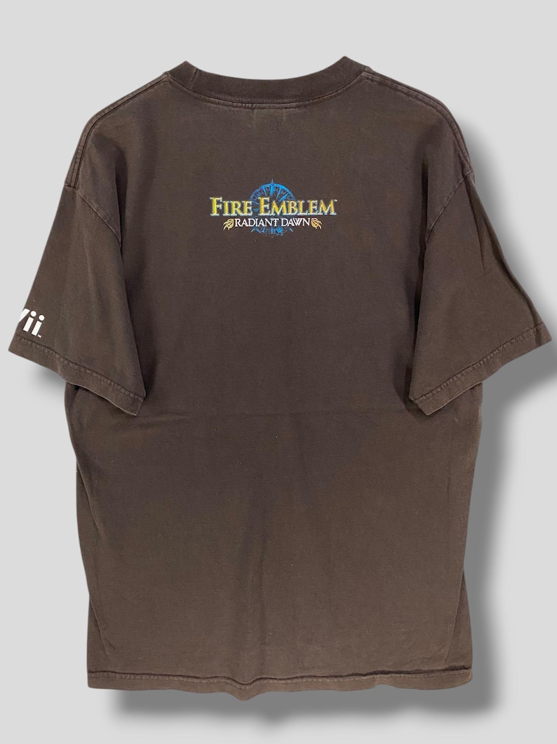 May include: Brown t-shirt with the text "Fire Emblem Radiant Dawn" in gold and blue. The shirt has a crew neck and short sleeves. The Wii logo is visible on the left sleeve.