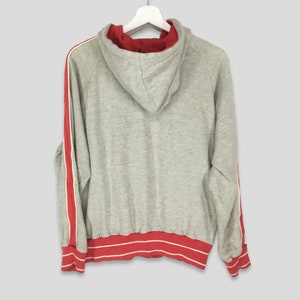 K&ouml;nnte beinhalten: Grauer Kapuzenpullover mit roten und wei&szlig;en Streifen an den &Auml;rmeln und am Saum. Die Innenseite der Kapuze ist rot. Der Pullover h&auml;ngt an einem wei&szlig;en Kleiderb&uuml;gel.