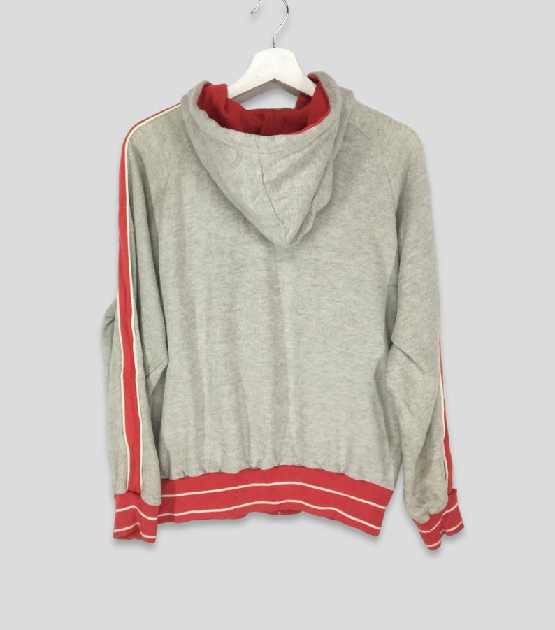 K&ouml;nnte beinhalten: Grauer Kapuzenpullover mit roten und wei&szlig;en Streifen an den &Auml;rmeln und am Saum. Die Innenseite der Kapuze ist rot. Der Pullover h&auml;ngt an einem wei&szlig;en Kleiderb&uuml;gel.