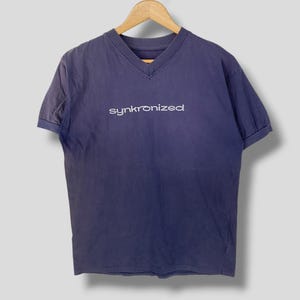 Puede incluir: Una camiseta azul oscuro con cuello en V y mangas cortas, que presenta la palabra synkronized impresa en blanco en el pecho. La prenda se muestra en una percha de madera, exhibiendo un diseño sencillo con cuello acanalado y mangas con dobladillo.