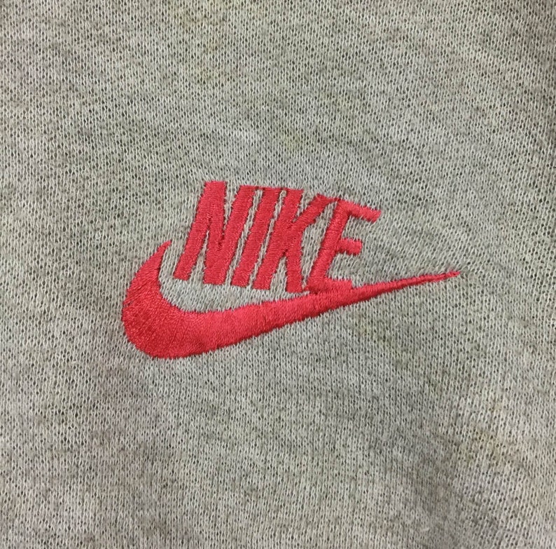 K&ouml;nnte beinhalten: Nahaufnahme eines grauen Stoffes mit einem roten Nike-Logo, das aufgestickt ist. Das Logo zeigt den Markennamen und das ikonische Swoosh-Design, ein klassisches Detail auf Sportbekleidung.