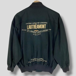 Könnte beinhalten: Eine dunkelgrüne Bomberjacke mit dem Text "LAUTREAMONT" in Gold auf der Rückseite gestickt. Der Text "ACRAFTSMAN AKARTISAN SPIRIT & SPONTANEOUSLY" befindet sich über dem Markennamen und "HOLIDAY SET A PARSONAT LIBERTYA HOLIDAY REISH" unter dem Markennamen. Der Text "LET'S TAKE A REST NOW" befindet sich unter der zweiten Textzeile. Der Text "EST ANARTISAN REAL LIVELY MODERN LEA AORT LIFE ONES AR OLD HOME ENCABURATE" ist unten auf der Jacke gestickt.