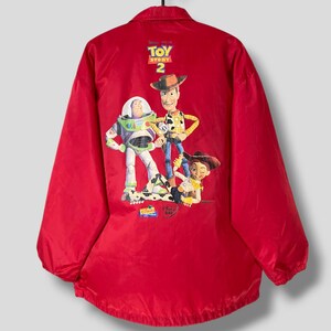 Puede incluir: Chaqueta de nylon roja con un gráfico de Toy Story 2 en la espalda. El gráfico presenta a Woody, Buzz Lightyear y Jessie. El texto "Toy Story 2" está impreso sobre los personajes.