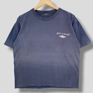 May include: A faded blue t-shirt with the text "RIP CURL" printed in white on the upper chest. The shirt is hanging on a wooden hanger. The t-shirt has short sleeves and a crew neck.