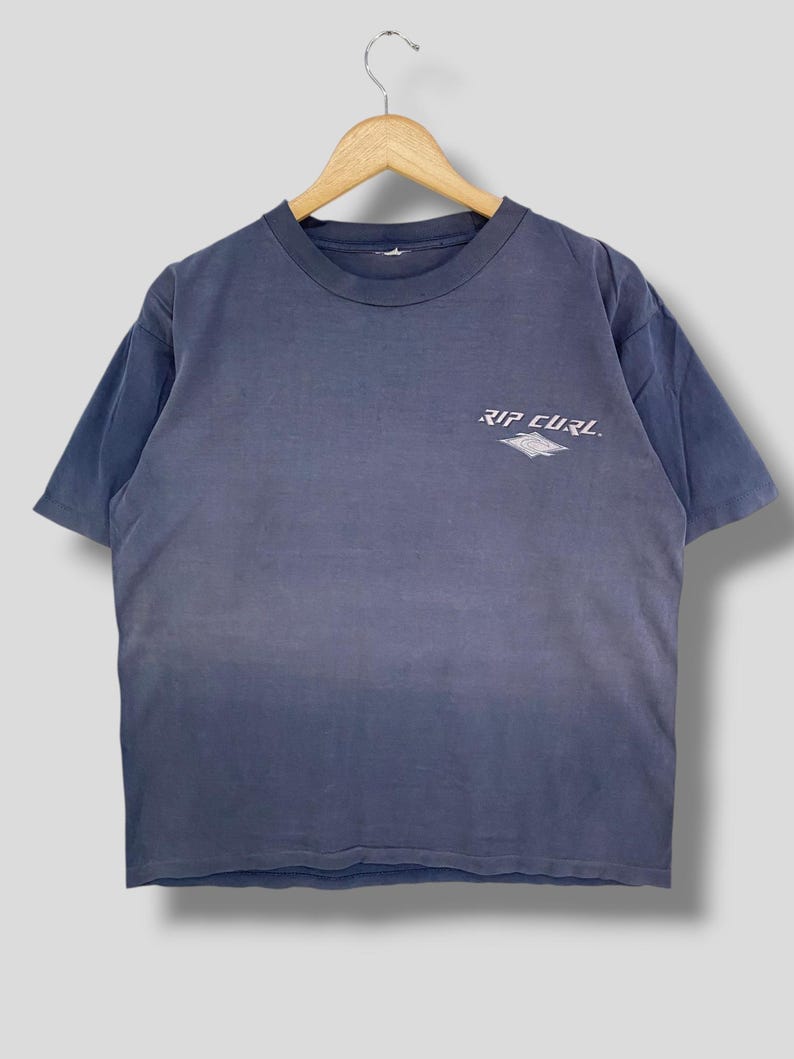 May include: A faded blue t-shirt with the text "RIP CURL" printed in white on the upper chest. The shirt is hanging on a wooden hanger. The t-shirt has short sleeves and a crew neck.
