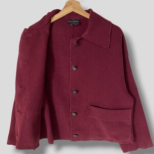 Puede incluir: Una chaqueta de lana color burdeos con cierre de botones y un solo bolsillo de parche en el lado izquierdo. La chaqueta tiene cuello y mangas largas. La etiqueta "McQ Alexander McQueen" es visible en el interior del cuello.