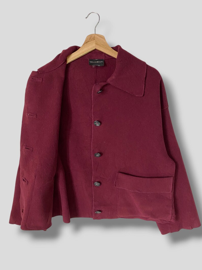 Puede incluir: Una chaqueta de lana color burdeos con cierre de botones y un solo bolsillo de parche en el lado izquierdo. La chaqueta tiene cuello y mangas largas. La etiqueta "McQ Alexander McQueen" es visible en el interior del cuello.