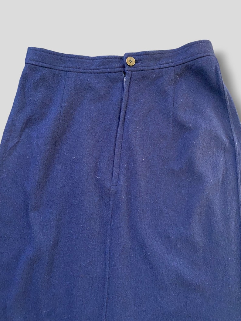 Puede incluir: Primer plano de una falda azul marino. La falda tiene un cierre de bot&oacute;n en la cintura y una costura vertical en la parte delantera. La tela parece ser una mezcla de lana. La falda es de color liso y tiene un dise&ntilde;o cl&aacute;sico.