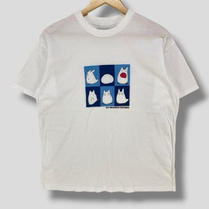 May include: White t-shirt featuring a graphic of characters from "My Neighbor Totoro" in a blue and white grid. The design includes various Totoro-like creatures. The text "MY NEIGHBOR TOTORO" is printed below the grid. The shirt is hanging on a wooden hanger.