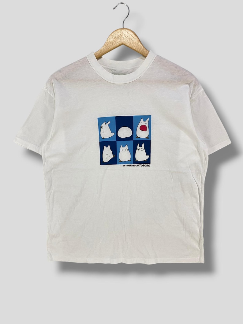 May include: White t-shirt featuring a graphic of characters from "My Neighbor Totoro" in a blue and white grid. The design includes various Totoro-like creatures. The text "MY NEIGHBOR TOTORO" is printed below the grid. The shirt is hanging on a wooden hanger.