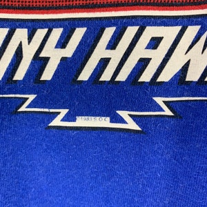 May include: A close-up of a blue t-shirt featuring the name "TONY HAWK" in large white letters. Below the name is a white lightning bolt design. The text "1983 S.O.C." is printed below the lightning bolt. The top of the shirt has a red and black striped design.