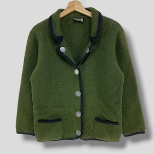 Könnte beinhalten: Grüne Wolljacke mit schwarzem Besatz und silbernen Knöpfen. Die Jacke hat zwei Vordertaschen und hängt an einem Holzbügel. Der Kragen und die Taschen sind mit schwarzem Besatz umrandet.