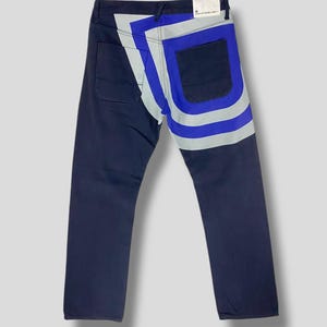 Puede incluir: Vaqueros azul marino oscuro con un diseño único en los bolsillos traseros. El diseño presenta una combinación de formas geométricas azules, plateadas y blancas. Los vaqueros tienen un diseño clásico de cinco bolsillos y corte recto.