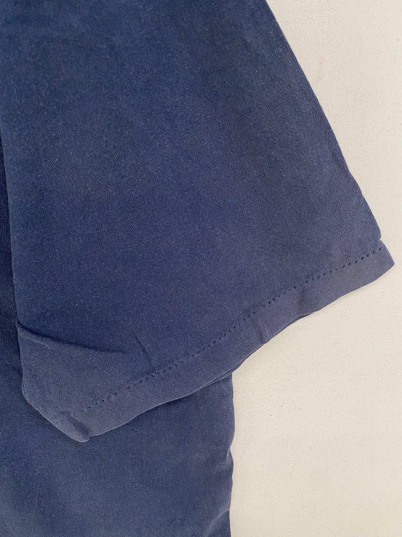 May include: Close-up of a navy blue t-shirt sleeve. The fabric is a solid color with a visible seam at the hem of the sleeve. The sleeve is slightly folded, showing the inside of the fabric.
