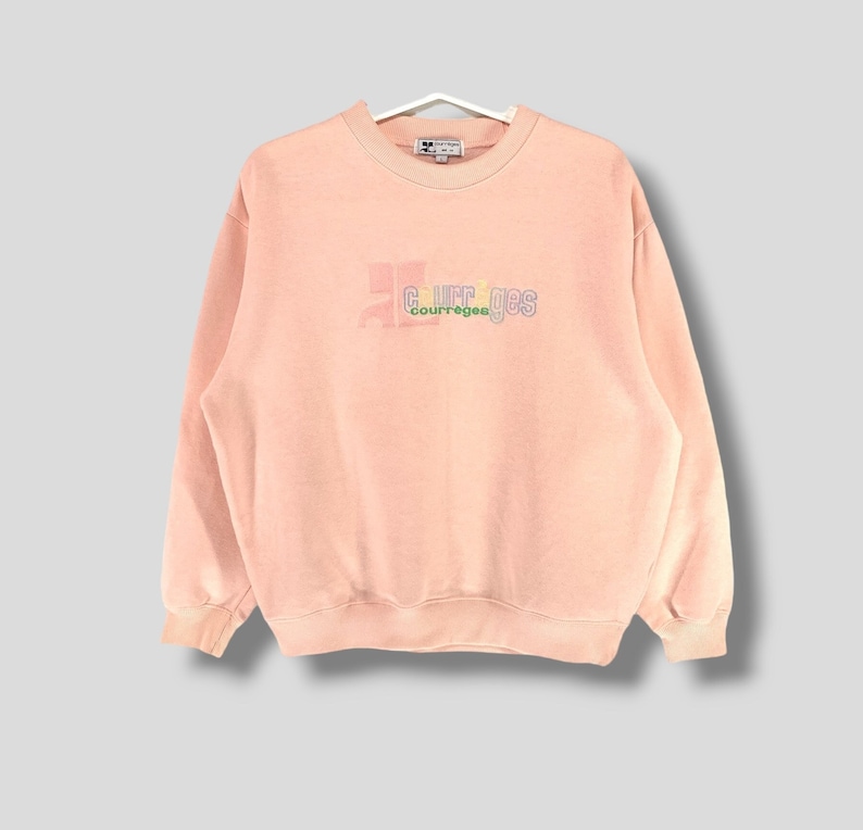 May include: A light pink Courr&egrave;ges sweatshirt with a crew neck and long sleeves. The brand name is embroidered on the front in a multi-colored design. The sweatshirt is hanging on a white hanger.