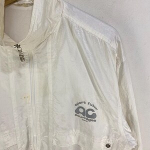 K&ouml;nnte beinhalten: Eine wei&szlig;e, leichte Jacke mit durchgehendem Rei&szlig;verschluss und hohem Kragen. Auf der Vorderseite der Jacke steht in Grau "sport futur ac courr&egrave;ges paris". Die Jacke h&auml;ngt an einem Holzkleiderb&uuml;gel.