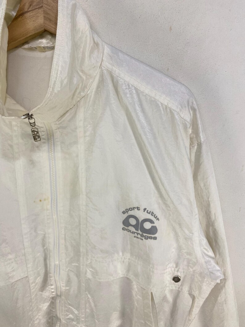 K&ouml;nnte beinhalten: Eine wei&szlig;e, leichte Jacke mit durchgehendem Rei&szlig;verschluss und hohem Kragen. Auf der Vorderseite der Jacke steht in Grau "sport futur ac courr&egrave;ges paris". Die Jacke h&auml;ngt an einem Holzkleiderb&uuml;gel.