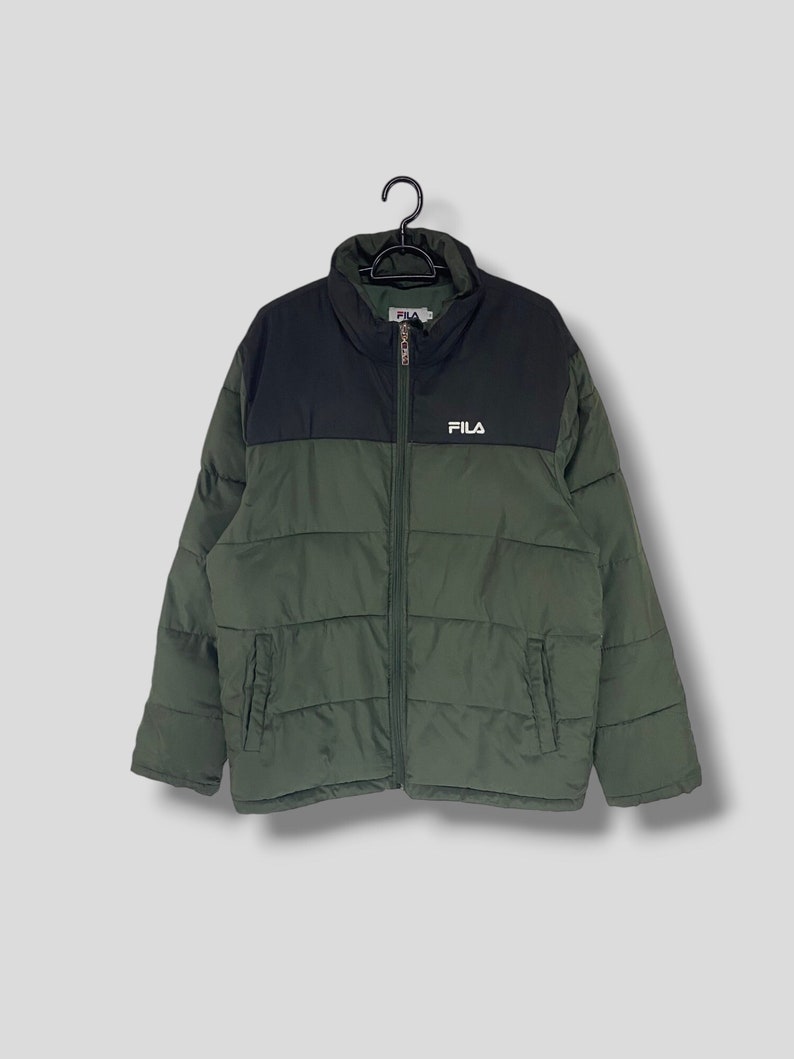 May include: A green and black FILA puffer jacket hanging on a black hanger. The jacket has a high collar and a full-length zipper. The FILA logo is displayed on the chest. The jacket is a solid color with a quilted design.