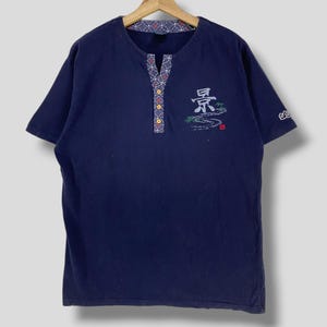 May include: Navy blue short-sleeved shirt with a V-neck and decorative trim. The shirt features embroidered Japanese characters and a landscape design. The shirt is hanging on a wooden hanger.
