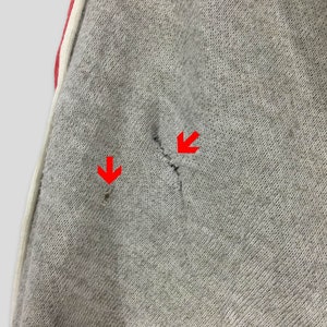 K&ouml;nnte beinhalten: Nahaufnahme einer grauen Jogginghose mit rotem Streifen und wei&szlig;em Besatz. Der Stoff hat zwei kleine L&ouml;cher und einen Riss. Die Jogginghose ist aus einem weichen, strukturierten Material gefertigt.