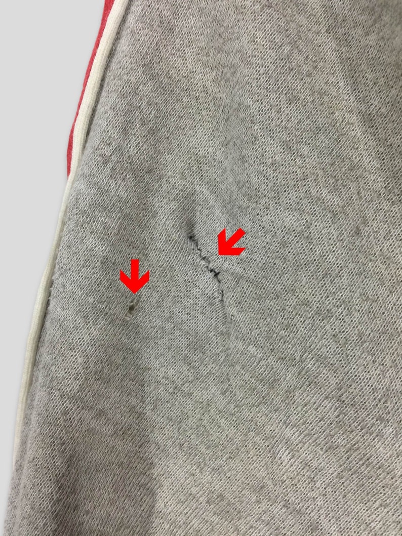 K&ouml;nnte beinhalten: Nahaufnahme einer grauen Jogginghose mit rotem Streifen und wei&szlig;em Besatz. Der Stoff hat zwei kleine L&ouml;cher und einen Riss. Die Jogginghose ist aus einem weichen, strukturierten Material gefertigt.