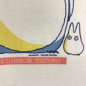 K&ouml;nnte beinhalten: Wei&szlig;es T-Shirt mit einer Grafik der Zeichentrickfigur Totoro, mit blauen und gelben Akzenten. Der Text "EIGHBOR TOTORO" ist auf einem pfirsichfarbenen Banner aufgedruckt. Das Design enth&auml;lt den Text "&copy;NIBARIKI &bull; TOKUMA SHOTEN".