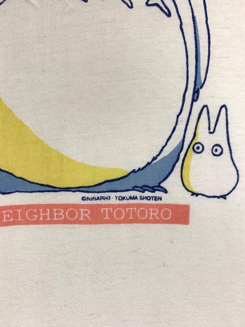 K&ouml;nnte beinhalten: Wei&szlig;es T-Shirt mit einer Grafik der Zeichentrickfigur Totoro, mit blauen und gelben Akzenten. Der Text "EIGHBOR TOTORO" ist auf einem pfirsichfarbenen Banner aufgedruckt. Das Design enth&auml;lt den Text "&copy;NIBARIKI &bull; TOKUMA SHOTEN".