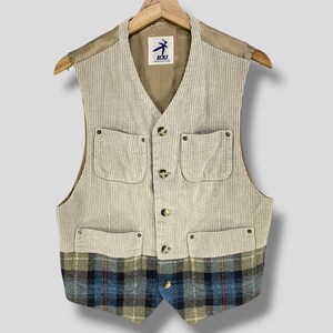 Peut inclure: Un gilet en velours côtelé beige avec une section inférieure à carreaux. Le gilet a quatre poches avant, une fermeture boutonnée et une doublure beige. Il est suspendu à un cintre en bois.