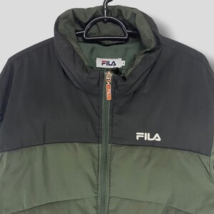 May include: A two-tone FILA puffer jacket in olive green and black. The jacket has a high collar, a full zipper, and the FILA logo in white on the chest. The jacket is hanging on a black hanger.