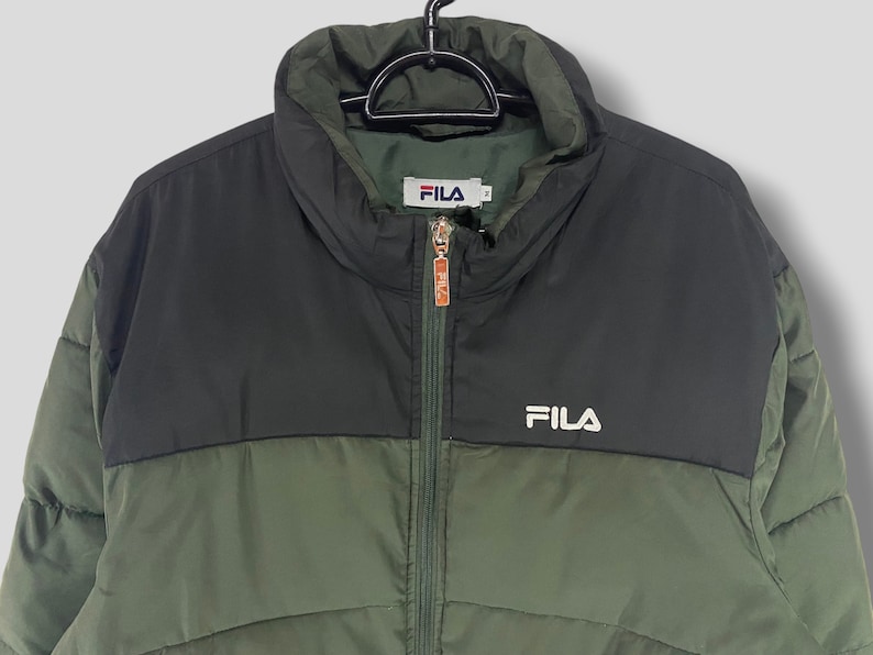 May include: A two-tone FILA puffer jacket in olive green and black. The jacket has a high collar, a full zipper, and the FILA logo in white on the chest. The jacket is hanging on a black hanger.