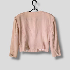 May include: A pale pink blazer with long sleeves and a button closure. The blazer has a fitted silhouette and is hanging on a black hanger.