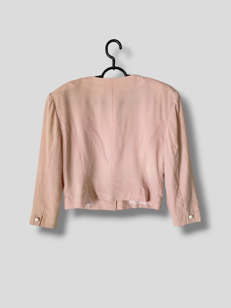 May include: A pale pink blazer with long sleeves and a button closure. The blazer has a fitted silhouette and is hanging on a black hanger.