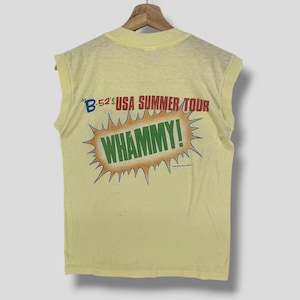 Könnte beinhalten: Ein ärmelloses, hellgelbes T-Shirt mit dem Text "The B-52's USA SUMMER TOUR WHAMMY!" in Blau, Rot und Grün. Die Grafik ist in einem Burst-Design gehalten. Das T-Shirt hängt an einem Holzbügel.