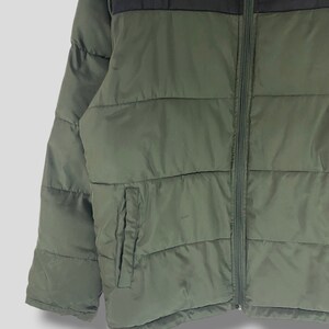 May include: Olive green and black puffer jacket with a full zipper closure. The jacket features a small pocket on the left side and the FILA logo in white on the upper right chest area. The jacket is designed with horizontal quilted panels.