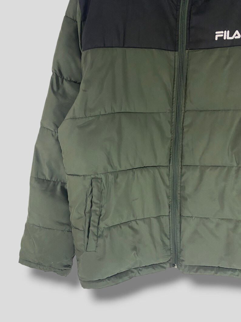 May include: Olive green and black puffer jacket with a full zipper closure. The jacket features a small pocket on the left side and the FILA logo in white on the upper right chest area. The jacket is designed with horizontal quilted panels.