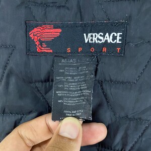 May include: A black Versace Sport label with white text. The label has a red logo with a stylized head of Medusa. The label includes information about the fabric and style of the garment, including the materials used and the country of origin. The label reads "Versace Sport", "Alias", "100% Polyamide/Polyamide", "Fodera/Lining", "100% Polyamide/Polyamide", "Imbottitura/Padding", "100% Polyester/Polyester", "Fabric and Style", "Made in Italy".