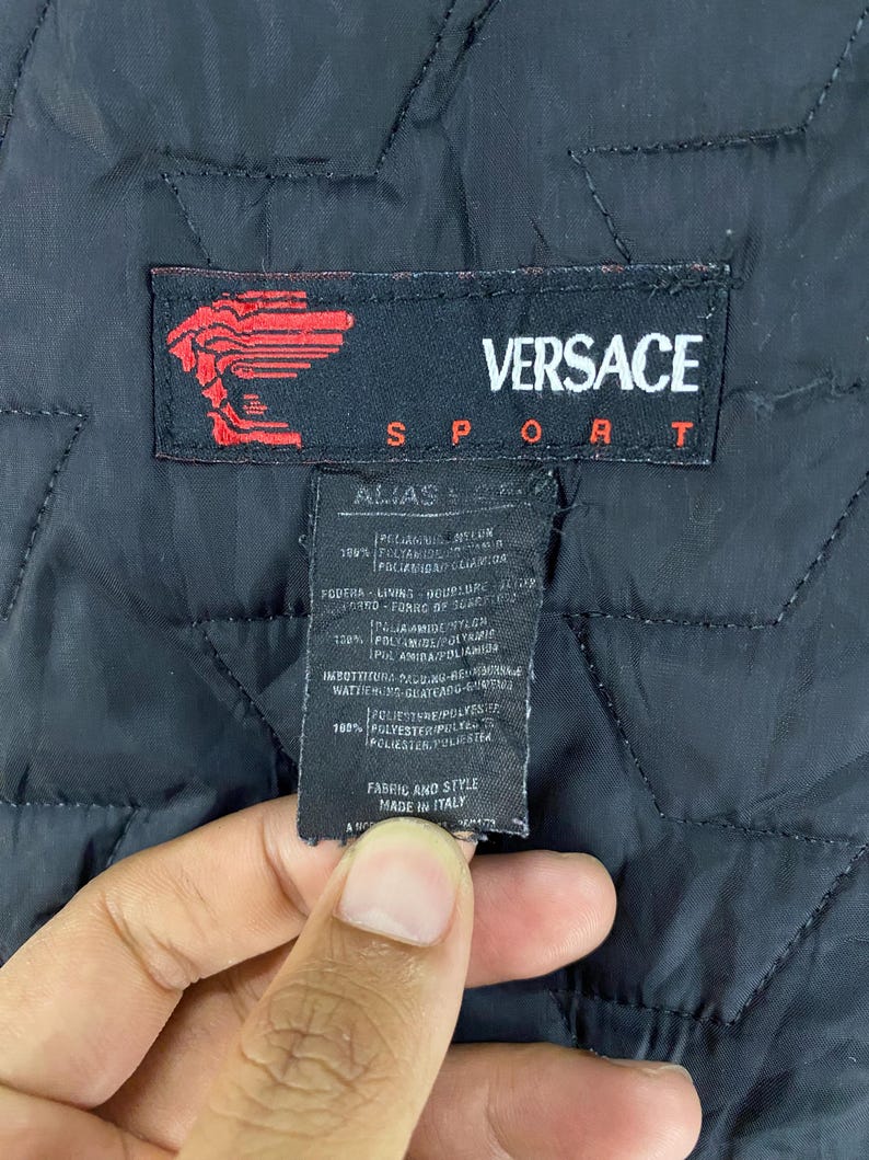 May include: A black Versace Sport label with white text. The label has a red logo with a stylized head of Medusa. The label includes information about the fabric and style of the garment, including the materials used and the country of origin. The label reads "Versace Sport", "Alias", "100% Polyamide/Polyamide", "Fodera/Lining", "100% Polyamide/Polyamide", "Imbottitura/Padding", "100% Polyester/Polyester", "Fabric and Style", "Made in Italy".
