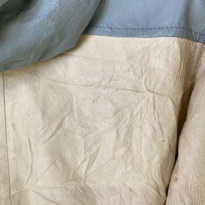 Puede incluir: Primer plano de una chaqueta de cuero azul claro y blanco roto. La chaqueta est&aacute; hecha de cuero suave y flexible y tiene una superficie texturizada.