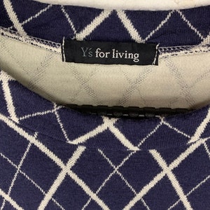 May include: Close-up of a navy blue and white argyle patterned sweater. The sweater features a diamond design and a black tag that reads "Y's for living". The sweater is made of a soft, knit fabric.