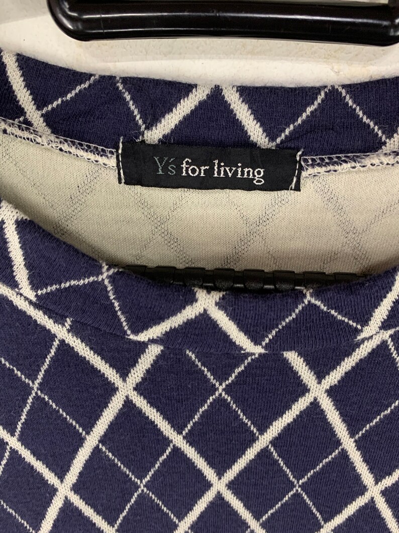 May include: Close-up of a navy blue and white argyle patterned sweater. The sweater features a diamond design and a black tag that reads "Y's for living". The sweater is made of a soft, knit fabric.