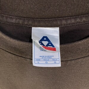 May include: Close-up of a brown t-shirt collar with a white tag. The tag has the letters "AA" in blue and red, with text in English and Spanish. The tag indicates the shirt is made in Mexico of USA fabric. Size L, G, and G are listed.