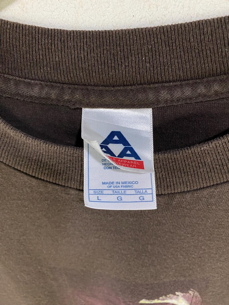 May include: Close-up of a brown t-shirt collar with a white tag. The tag has the letters "AA" in blue and red, with text in English and Spanish. The tag indicates the shirt is made in Mexico of USA fabric. Size L, G, and G are listed.