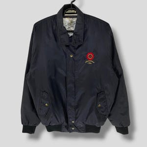 Op de afbeelding: Een zwarte bomberjack met een rood en gouden geborduurd logo met de tekst "International Star".