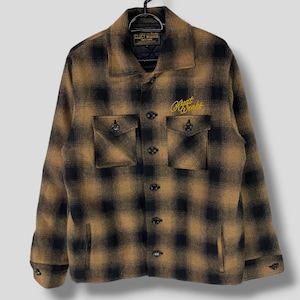 Puede incluir: Una camisa de franela a cuadros marrón y negro con un logotipo bordado en oro que dice "Blunt Worlds" en el pecho izquierdo. La camisa tiene un frente abotonado y dos bolsillos de solapa.