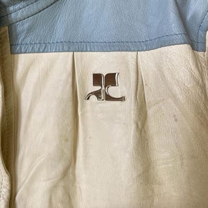 Puede incluir: Primer plano de una chaqueta de cuero blanca con cuello de cuero azul. La chaqueta tiene un logotipo de metal plateado en la parte delantera que dice "RC".