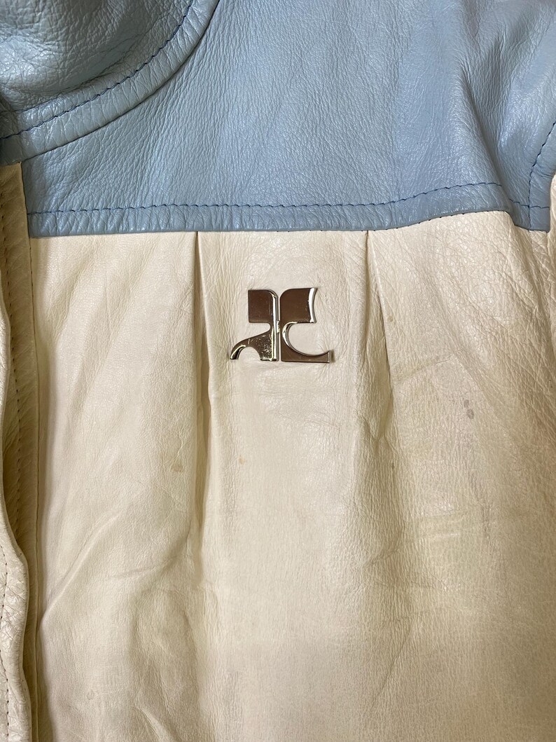 Puede incluir: Primer plano de una chaqueta de cuero blanca con cuello de cuero azul. La chaqueta tiene un logotipo de metal plateado en la parte delantera que dice "RC".