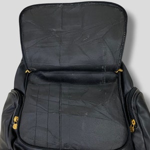 May include: Black leather backpack with an open top, revealing the interior compartments. The backpack features gold-colored zippers and white stitching details. The design includes multiple pockets and a structured shape, suitable for carrying various items.
